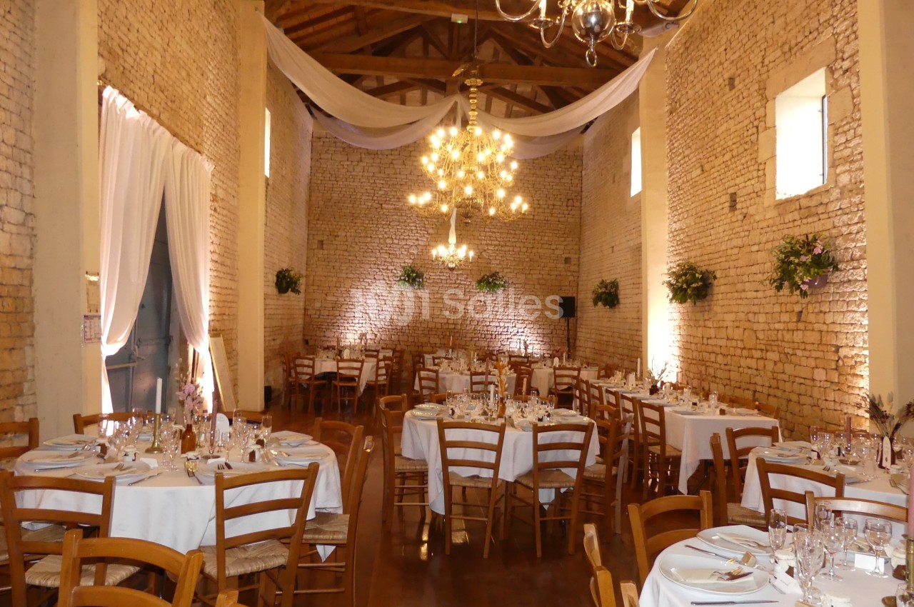 Salle de réception en pierre avec tables rondes dressées, chaises en bois, lustres suspendus et décorations florales.