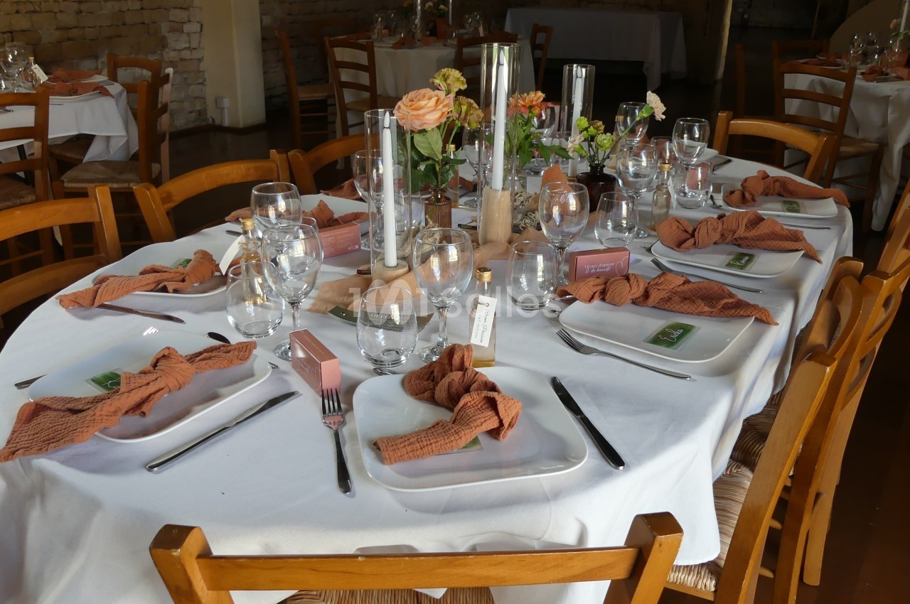 Table dressée avec nappes blanches, serviettes orange, verres, couverts et décorations florales dans un cadre chaleureux.