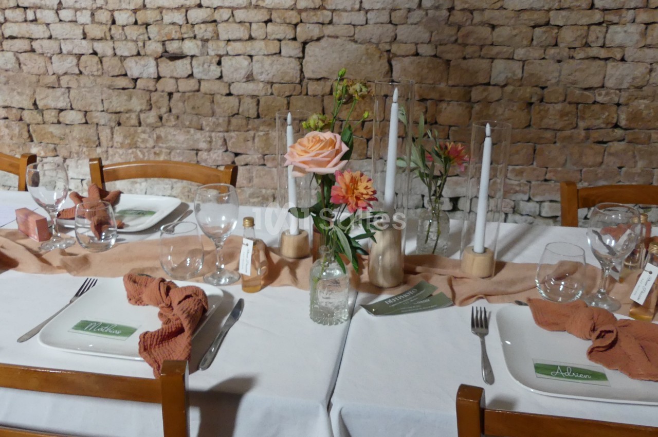 Table dressée avec nappes blanches, serviettes orange, fleurs, bougies et vaisselle devant un mur en pierre.