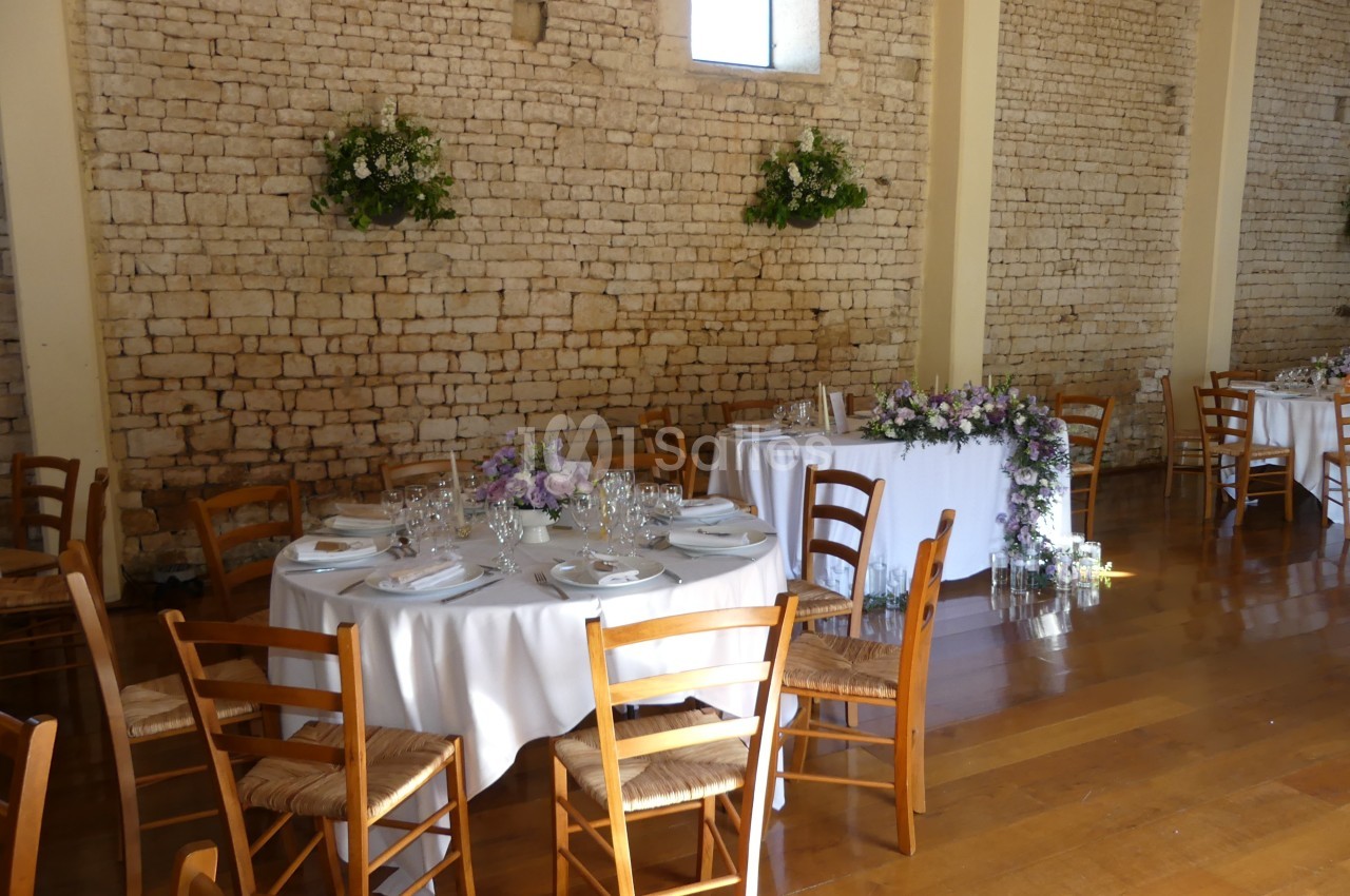 Salle de réception avec murs en pierre, tables rondes dressées, chaises en bois et décorations florales.