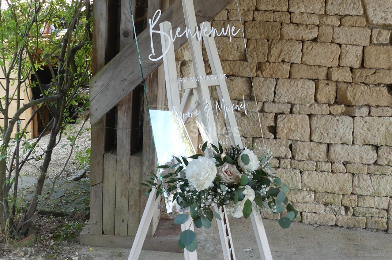 Panneau de bienvenue pour un mariage, en verre transparent, décoré de fleurs blanches et vertes, posé sur un chevalet.