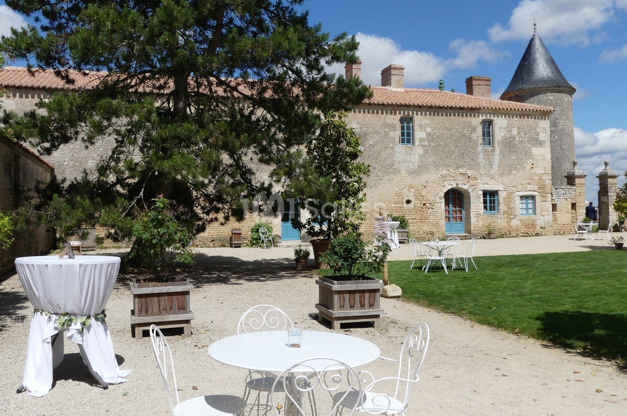 Cour d'un château avec mobilier de jardin blanc, table dressée et pelouse, sous un ciel partiellement nuageux.