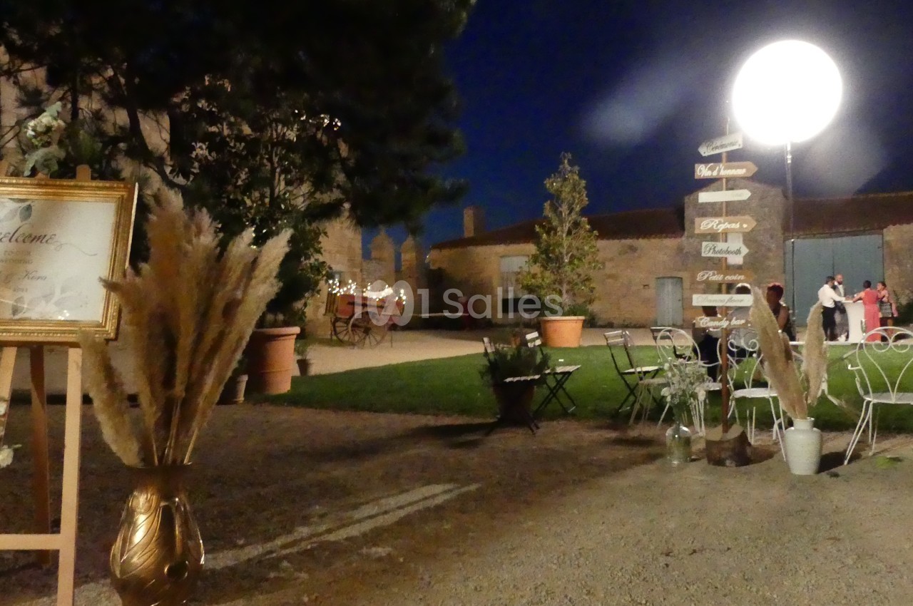 Espace extérieur éclairé de nuit avec des tables, des chaises, des décorations végétales et un panneau directionnel.