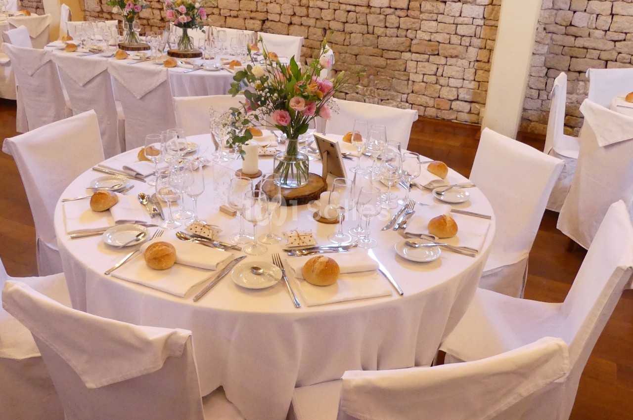 Table ronde élégamment dressée avec nappes blanches, vaisselle, fleurs au centre et chaises couvertes.