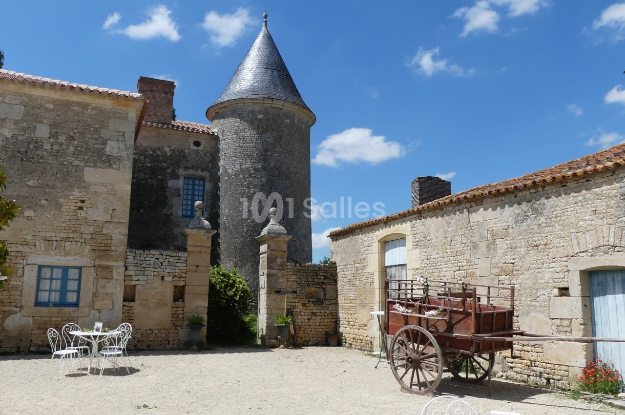 Cour intérieure d'un bâtiment en pierre avec une tour, une charrette en bois et des tables blanches sous un ciel bleu.