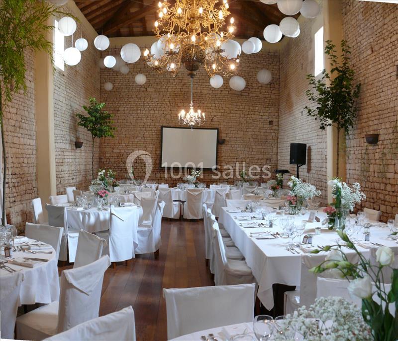 Salle de réception en pierre avec tables dressées, chaises blanches, décorations florales et lustres suspendus.