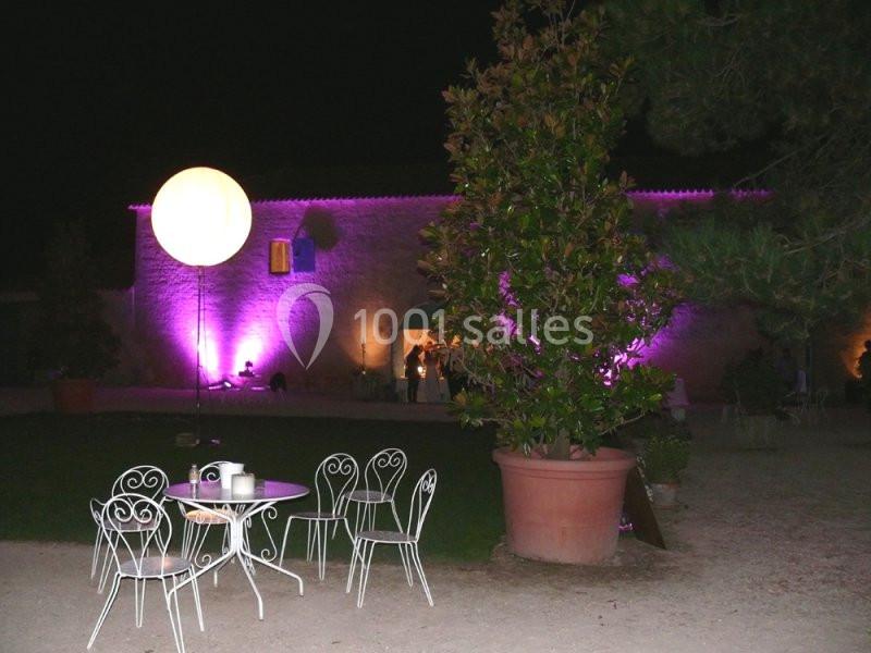 Cour extérieure éclairée de nuit avec des chaises blanches, une table et un bâtiment illuminé en arrière-plan.