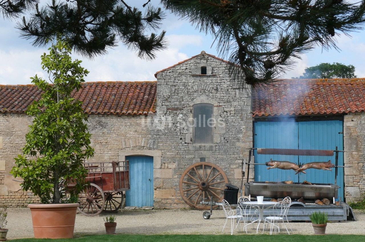 Cour d'une ferme avec murs en pierre, charrette ancienne, table en fer forgé et rôtissoire en fonctionnement.