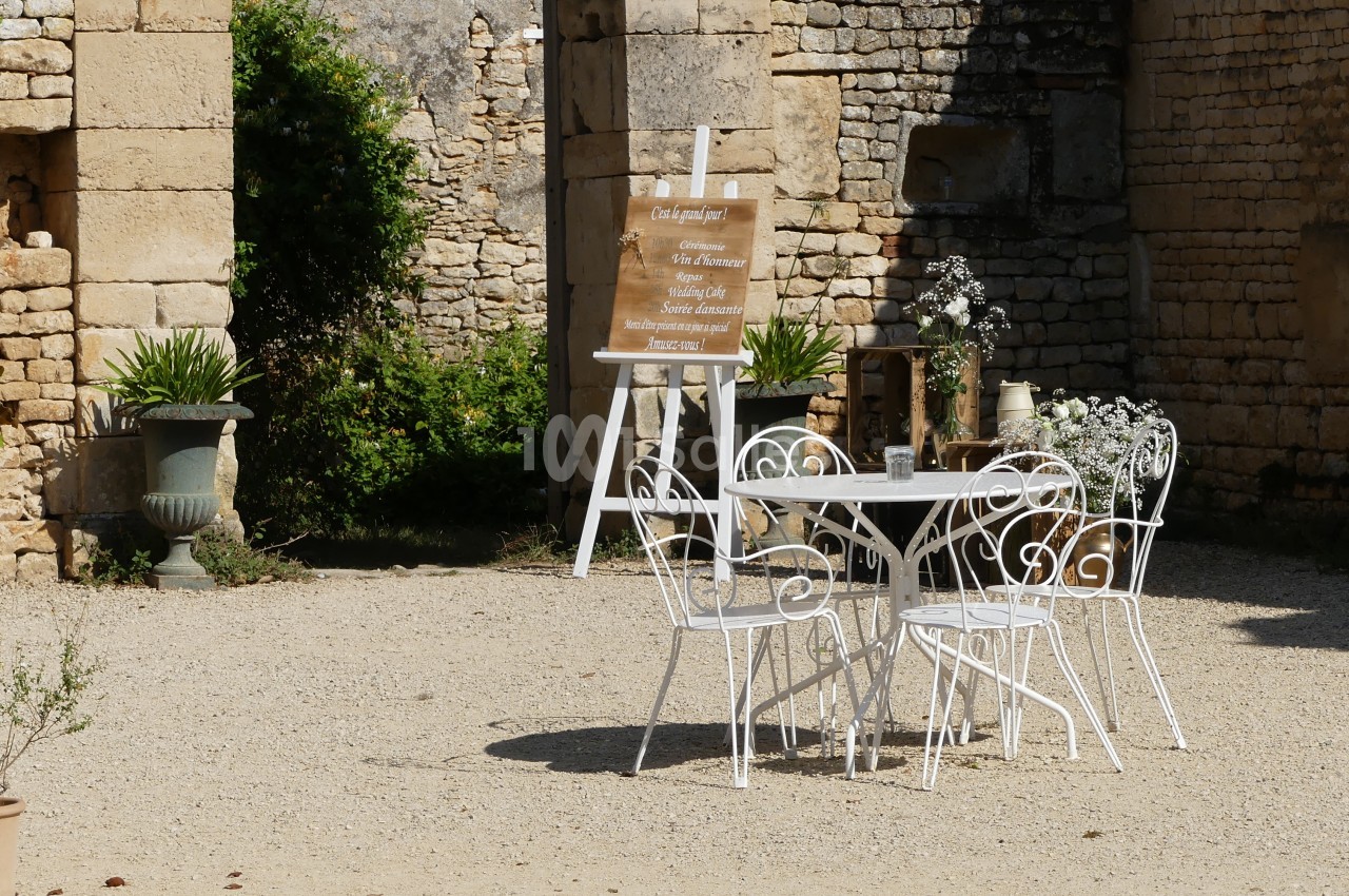 Table et chaises blanches en fer forgé disposées sur une terrasse en gravier, entourées de plantes et de murs en pierre.