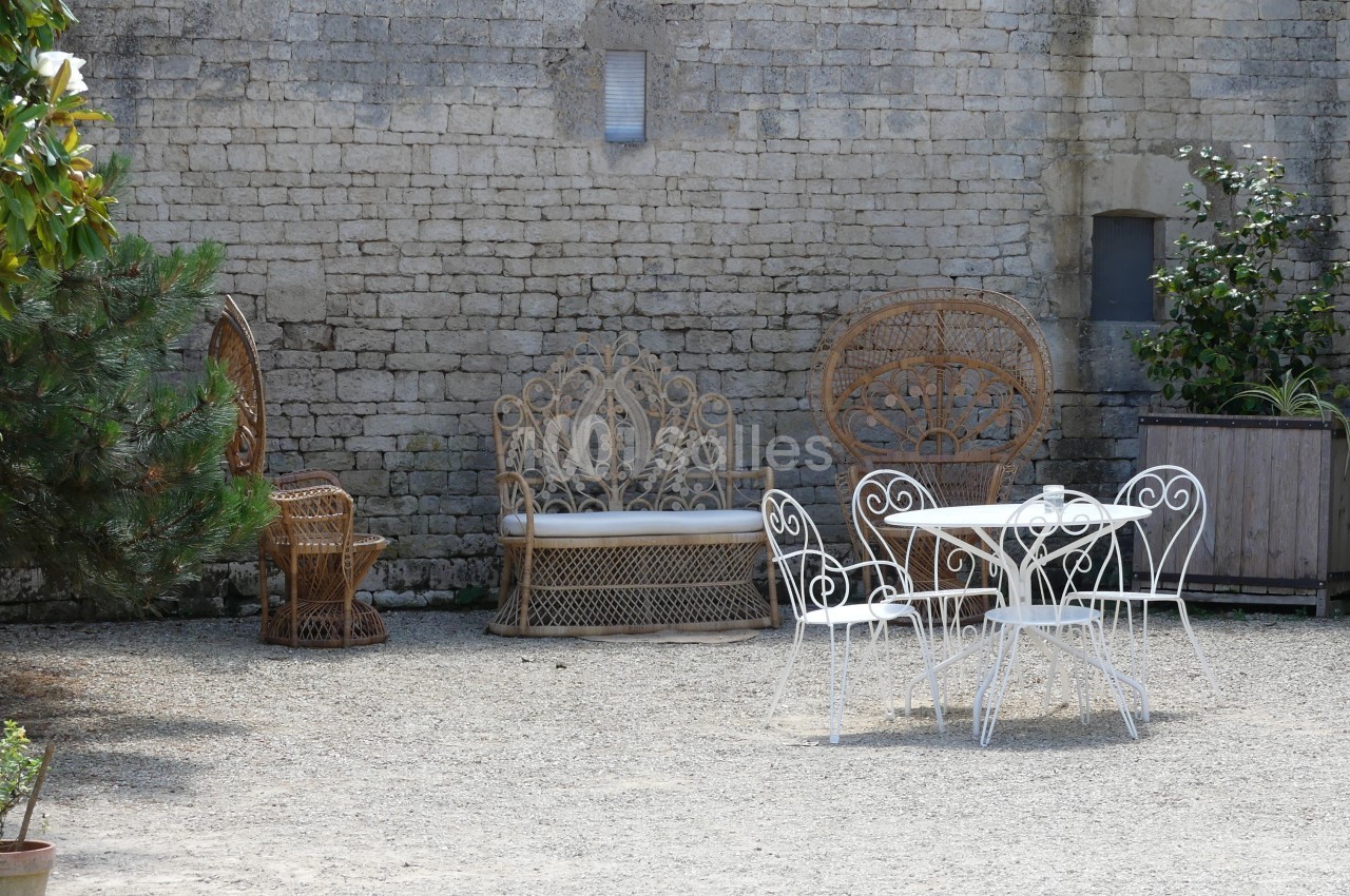 Mobilier de jardin en fer forgé et rotin disposé sur une cour en gravier devant un mur en pierre.