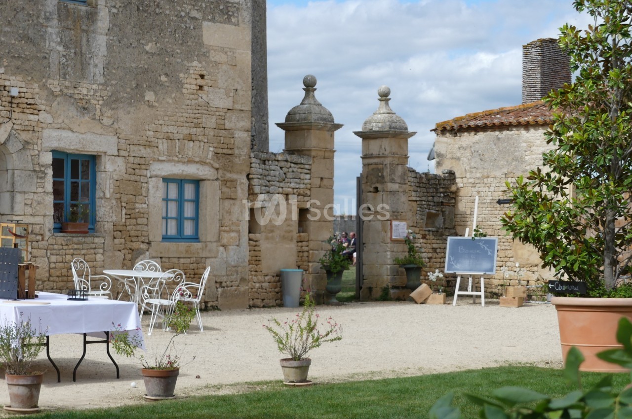 Cour d'une bâtisse en pierre avec mobilier de jardin, tableau noir et plantes en pot, sous un ciel partiellement nuageux.