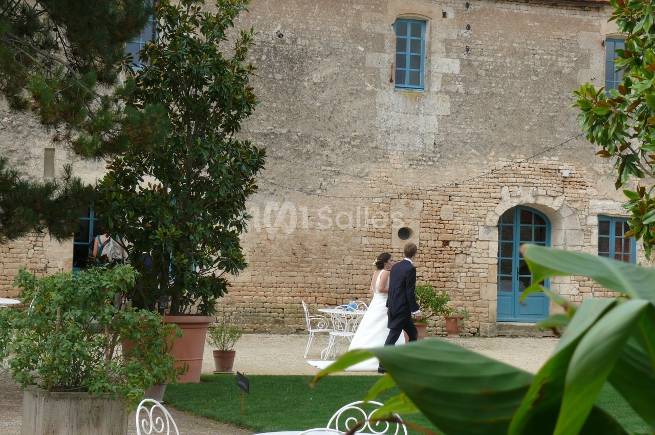 Un couple marche dans la cour d'une maison en pierre avec des fenêtres bleues, entourée de végétation.