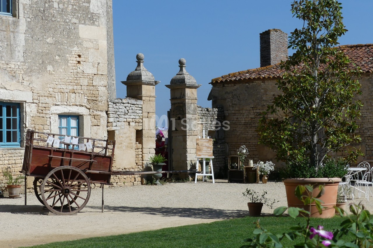 Cour d'une bâtisse en pierre avec une charrette en bois, des plantes en pot et des meubles de jardin sous un ciel bleu.