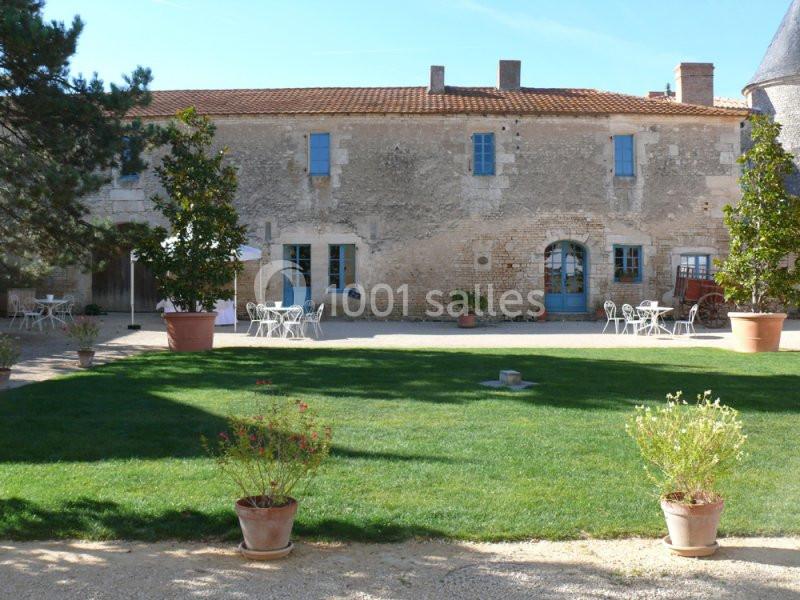 Cour d'une maison en pierre avec pelouse, tables et chaises extérieures, entourée de pots de fleurs et d'arbres.