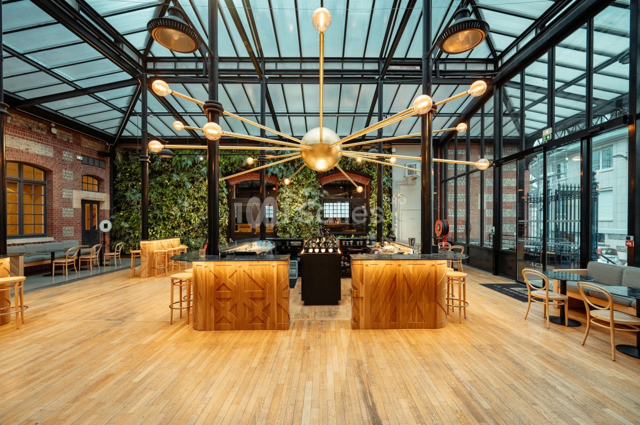 Intérieur d'un restaurant lumineux avec verrière, grande suspension dorée, murs végétalisés et mobilier en bois.