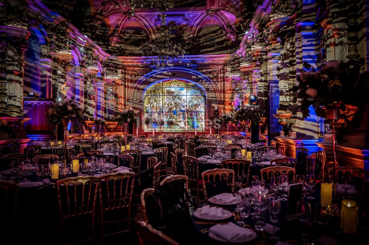 Salle de réception décorée avec des tables dressées, éclairages colorés et vitrail en arrière-plan.