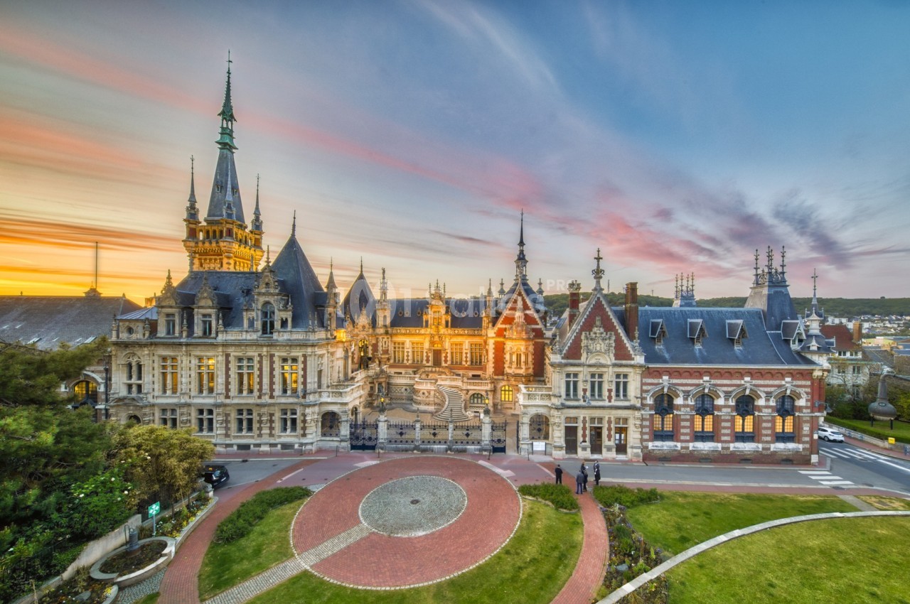 Vue d'un bâtiment historique avec une architecture détaillée, entouré de jardins, au coucher du soleil.