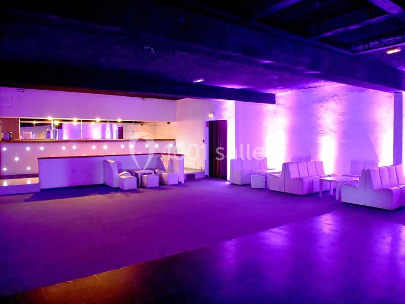 Salle vide avec éclairage violet, bar lumineux à gauche et banquettes blanches disposées le long des murs.