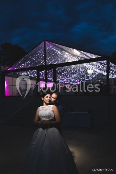 Un couple en tenue de mariage pose devant une structure éclairée par des guirlandes lumineuses sous un ciel nocturne.
