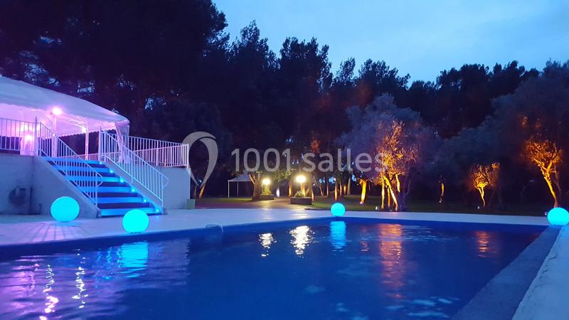 Piscine éclairée de nuit avec des lumières colorées et des arbres illuminés en arrière-plan.
