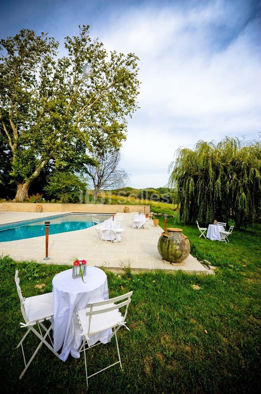 Tables blanches dressées près d'une piscine entourée de verdure et d'arbres sous un ciel légèrement nuageux.