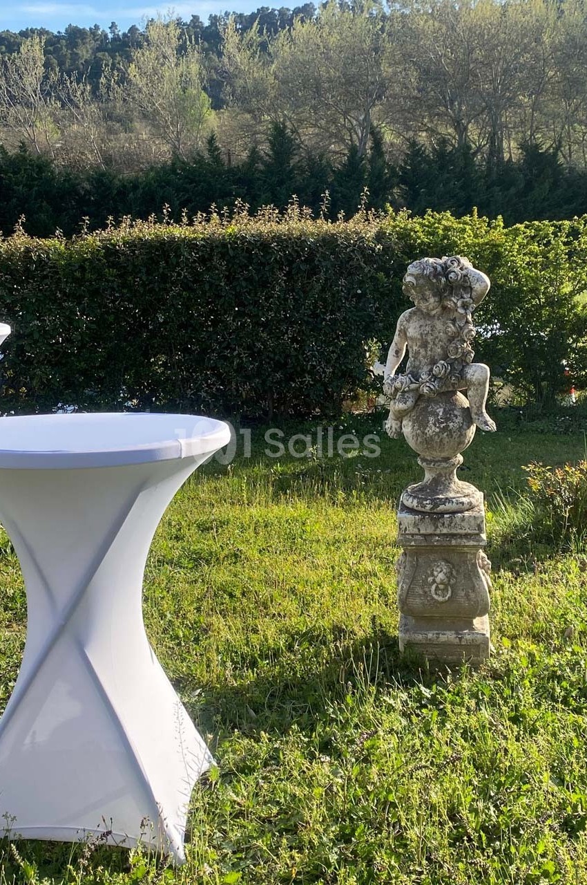 Statue en pierre représentant un enfant assis sur un piédestal, entourée de tables hautes et de verdure.