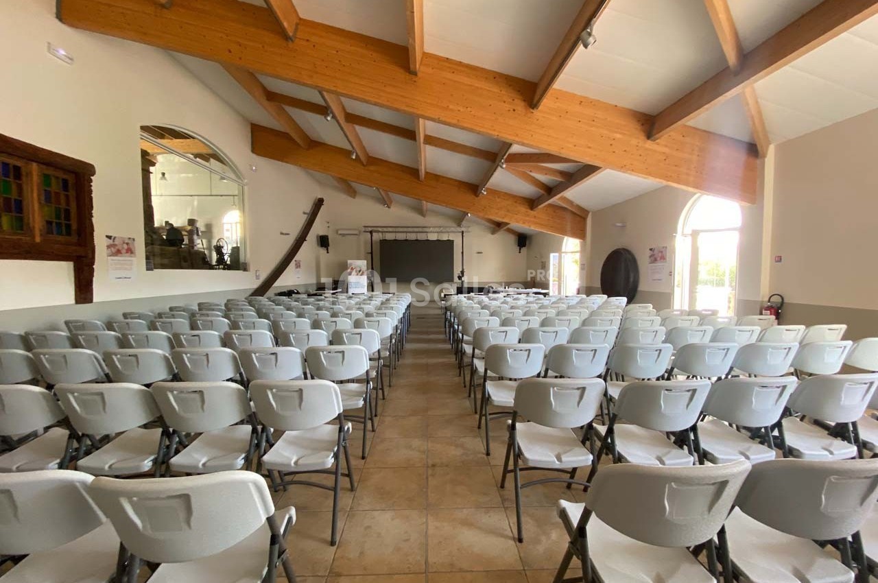 Salle lumineuse avec des rangées de chaises blanches alignées face à un écran et une scène, sous une charpente en bois.