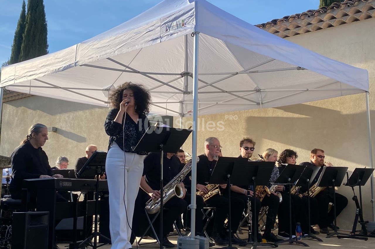 Chanteuse interprétant une chanson sur scène, accompagnée d'un orchestre sous une tente blanche en extérieur.