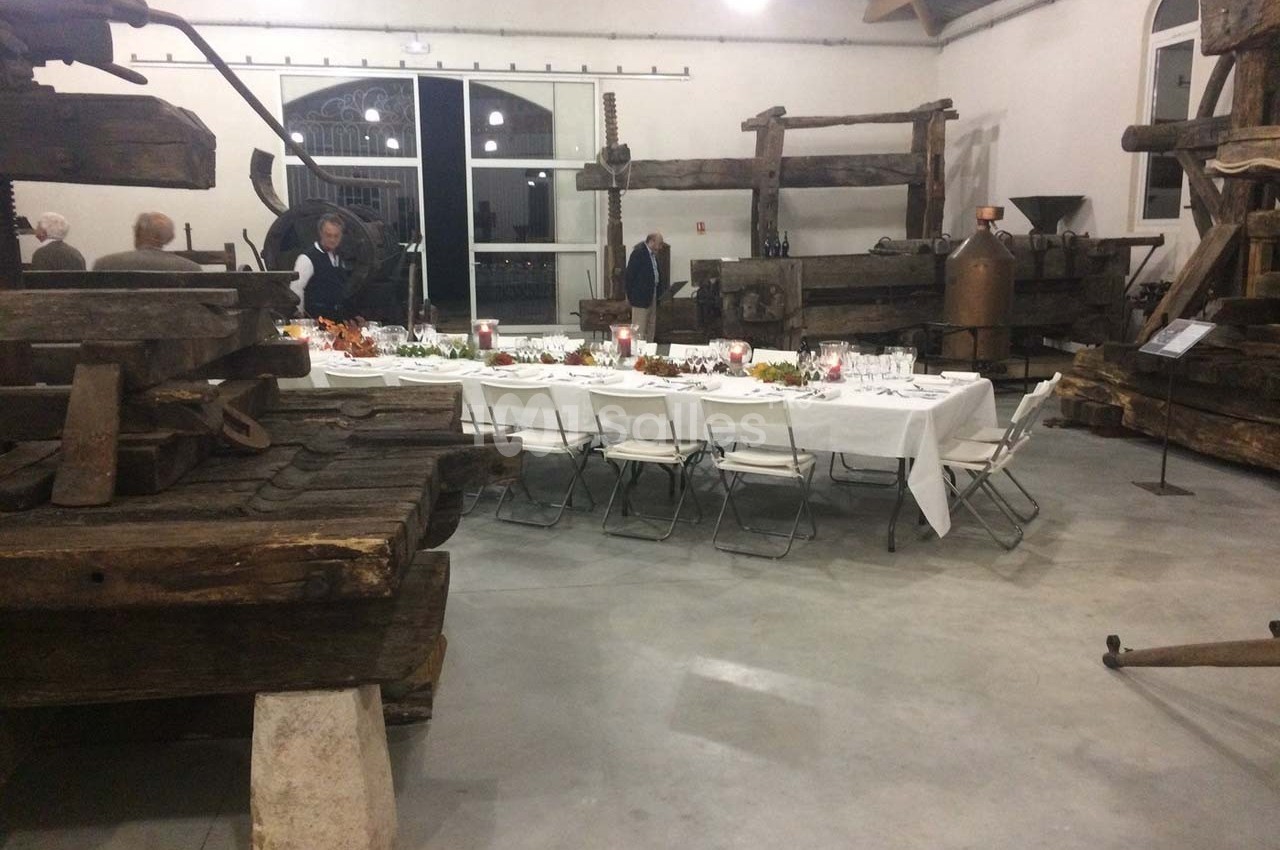 Salle avec des pressoirs anciens en bois, une longue table dressée pour un repas, et quelques personnes en arrière-plan.