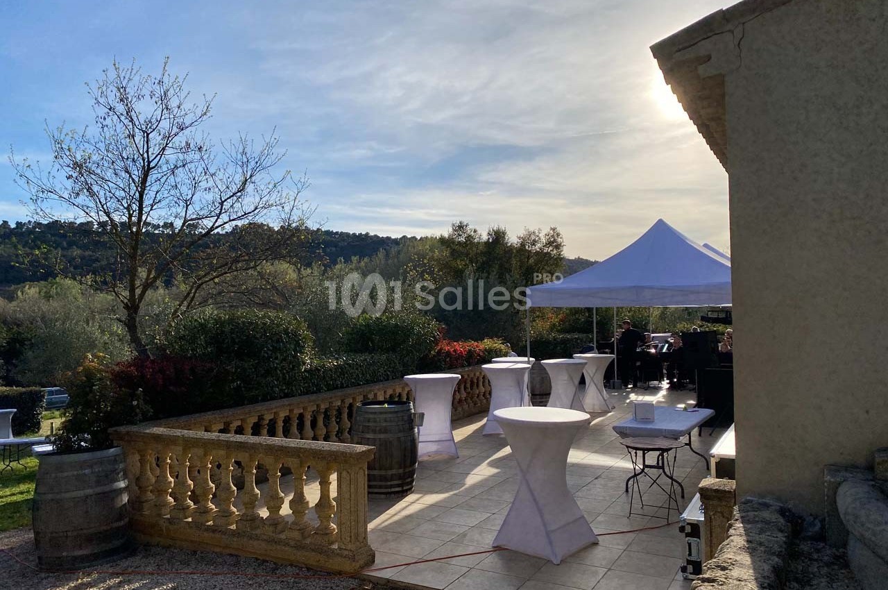 Terrasse ensoleillée avec tables hautes et tonnelle blanche, entourée de végétation et collines en arrière-plan.