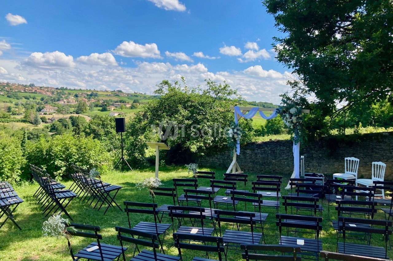 Chaises noires disposées en rangées sur une pelouse, face à une arche décorée, avec vue sur une campagne vallonnée.