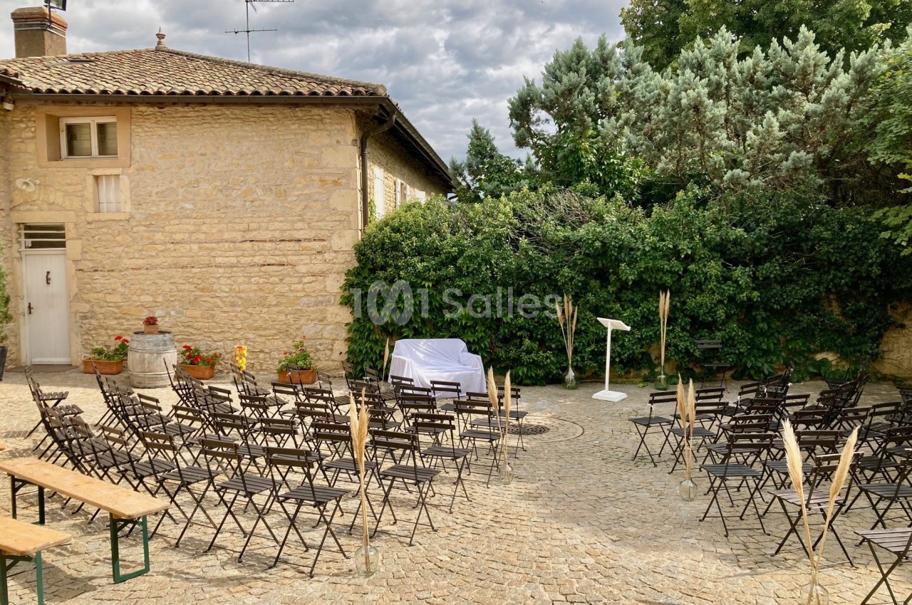Chaises et bancs disposés en extérieur sur une cour pavée, face à un pupitre blanc, entourés de verdure.