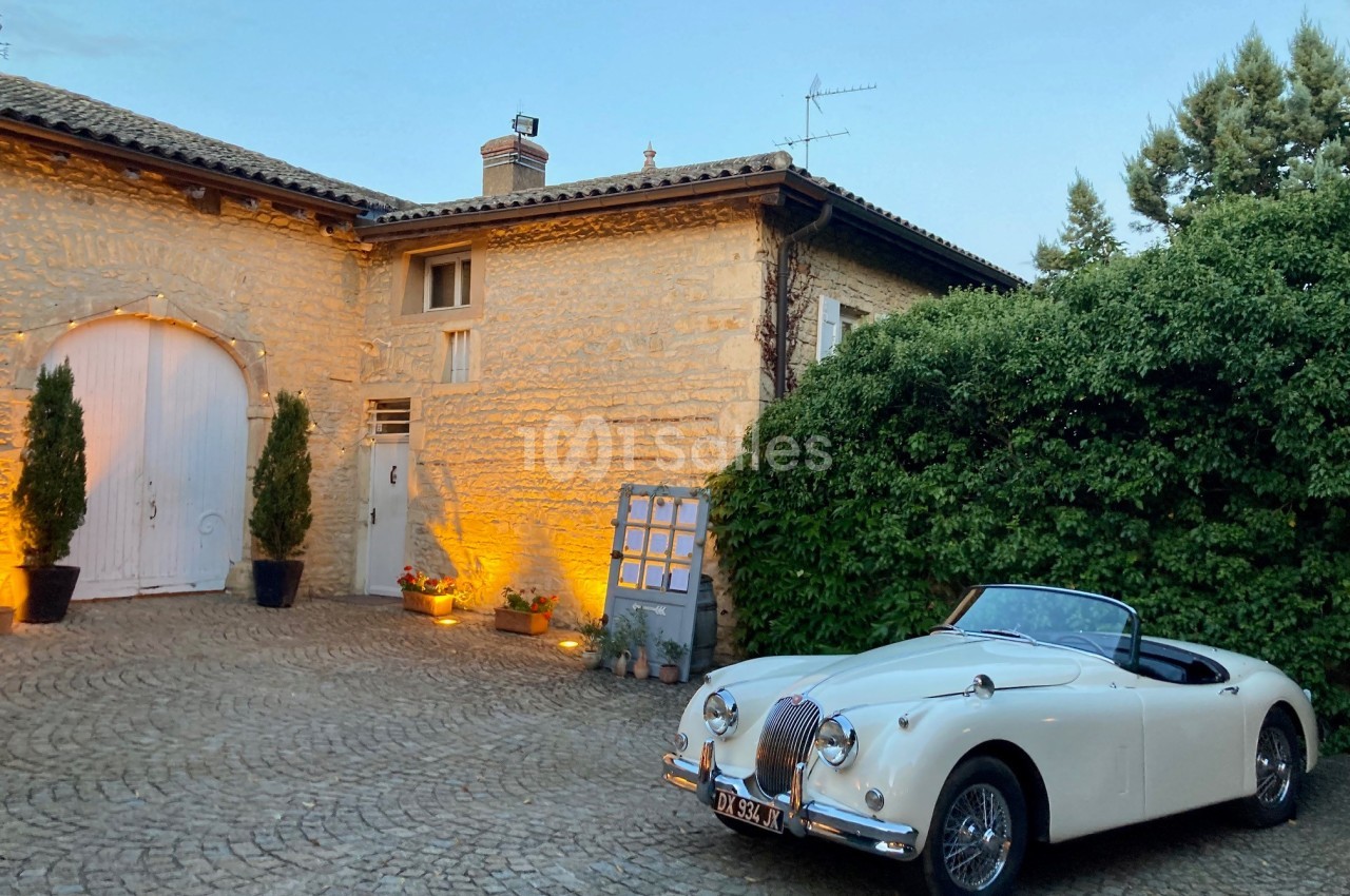 Voiture de collection blanche décapotable stationnée devant une maison en pierre avec éclairage chaleureux au crépuscule.