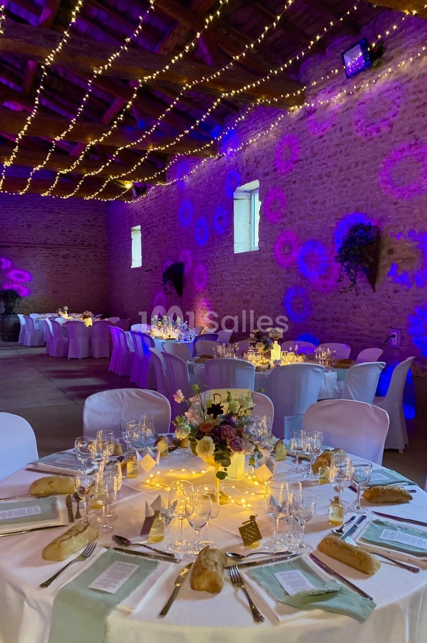 Salle de réception décorée avec des guirlandes lumineuses, tables rondes dressées et éclairage d'ambiance coloré.