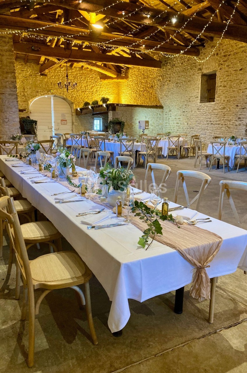 Salle de réception rustique avec tables décorées de nappes blanches, plantes et lumières suspendues au plafond en bois.
