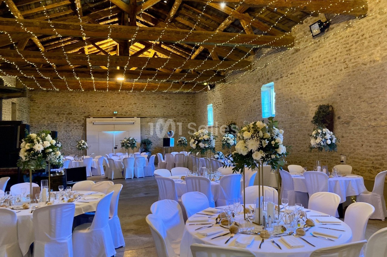 Salle de réception en pierre avec tables décorées de fleurs blanches, chaises blanches et guirlandes lumineuses au plafond.