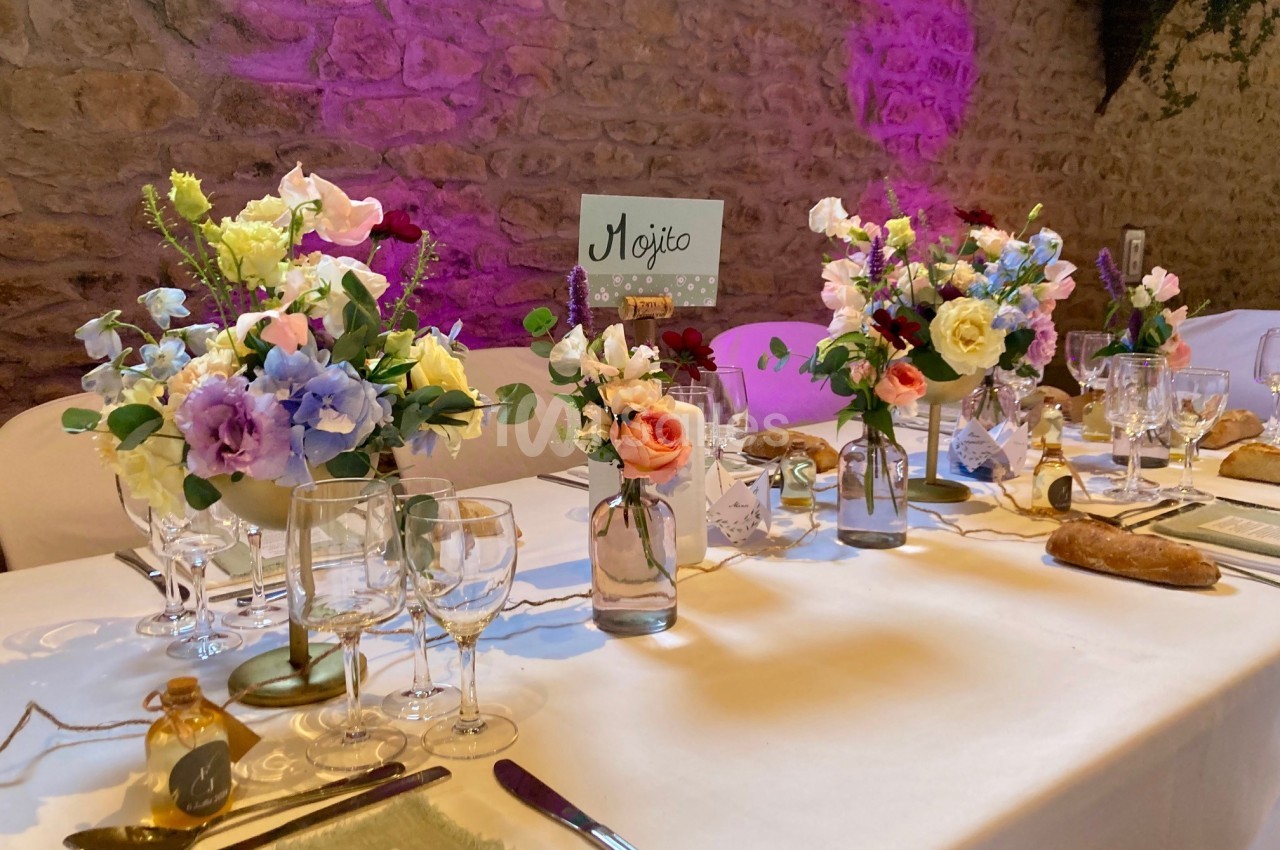 Table décorée pour un événement avec fleurs colorées, vaisselle élégante et pancarte ’Mojito’ sur fond de mur en pierre.