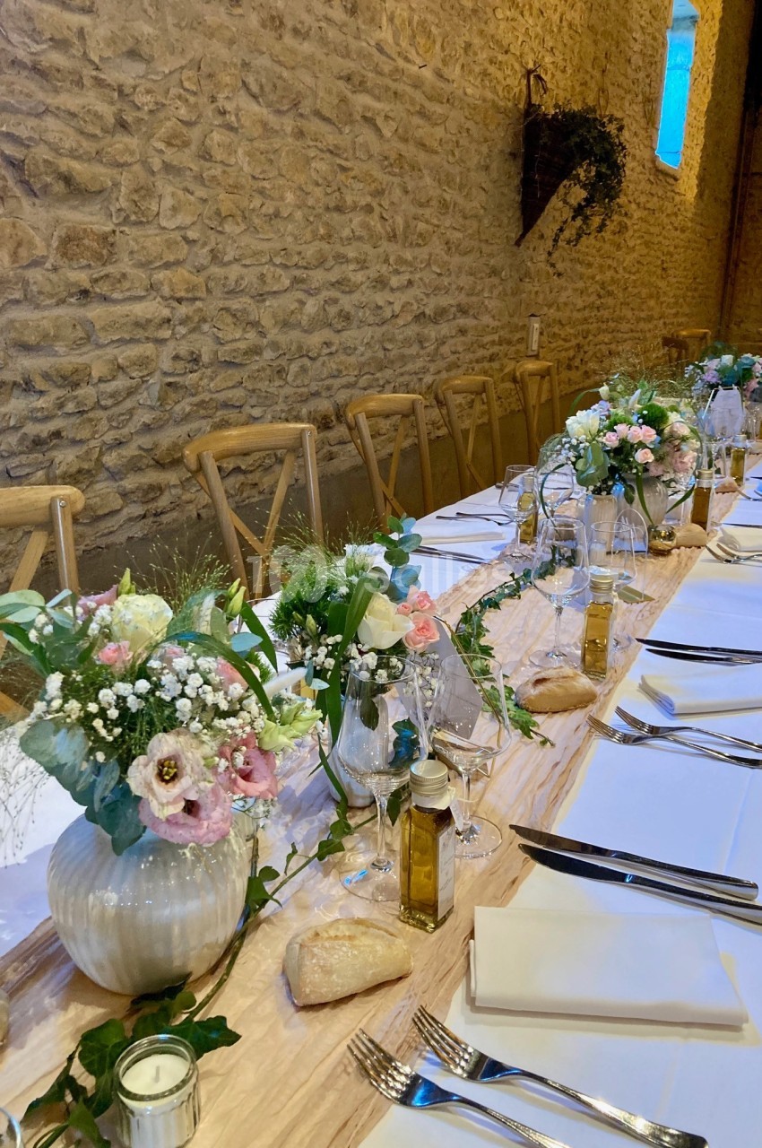 Table de réception décorée avec des fleurs, nappes blanches, vaisselle élégante et murs en pierre apparente.