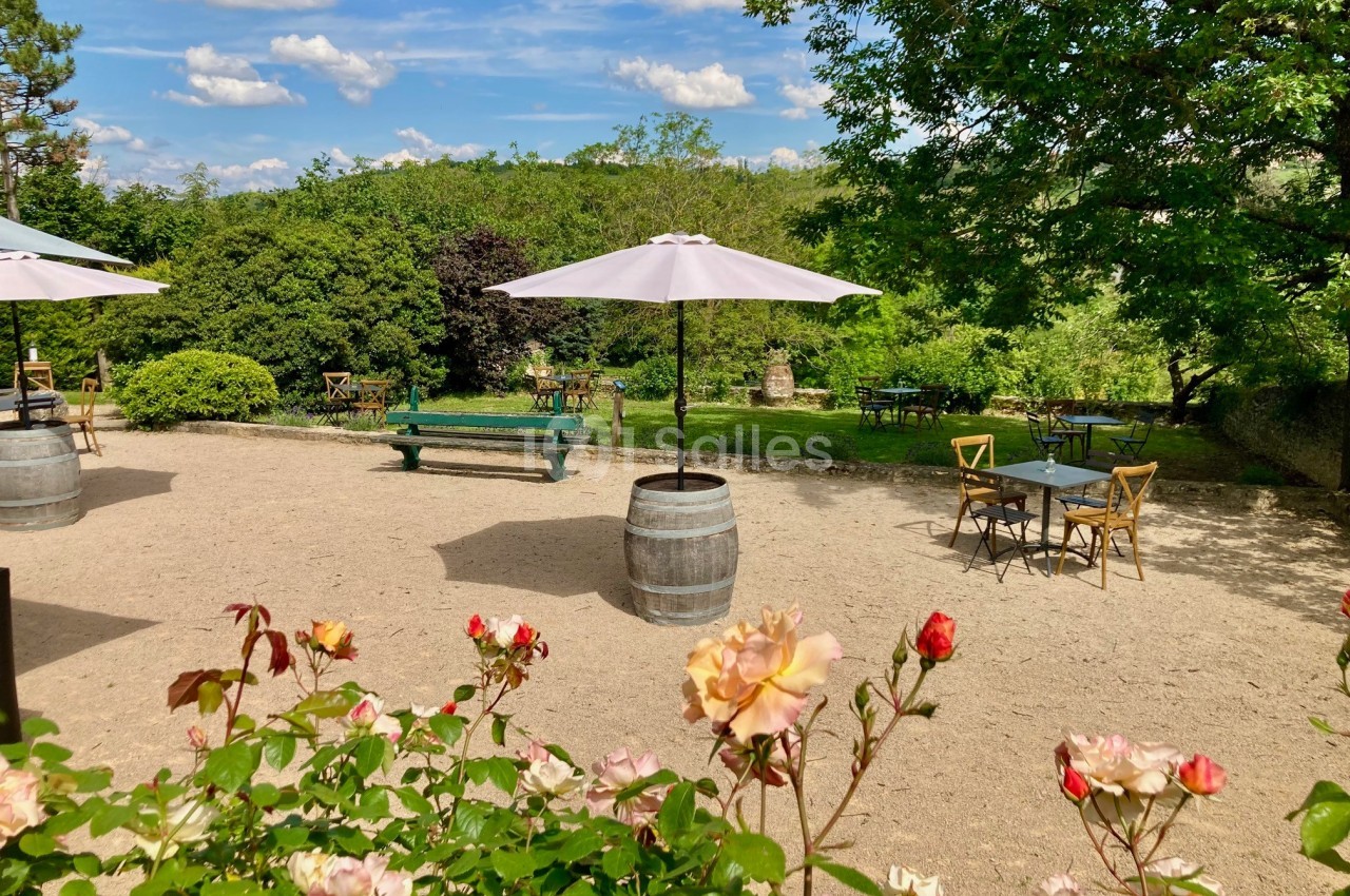 Terrasse extérieure avec tables, chaises et parasols, entourée de verdure et de fleurs sous un ciel ensoleillé.