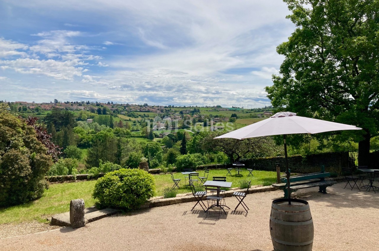 Terrasse avec tables et parasol, offrant une vue dégagée sur un paysage vallonné et verdoyant sous un ciel partiellement…