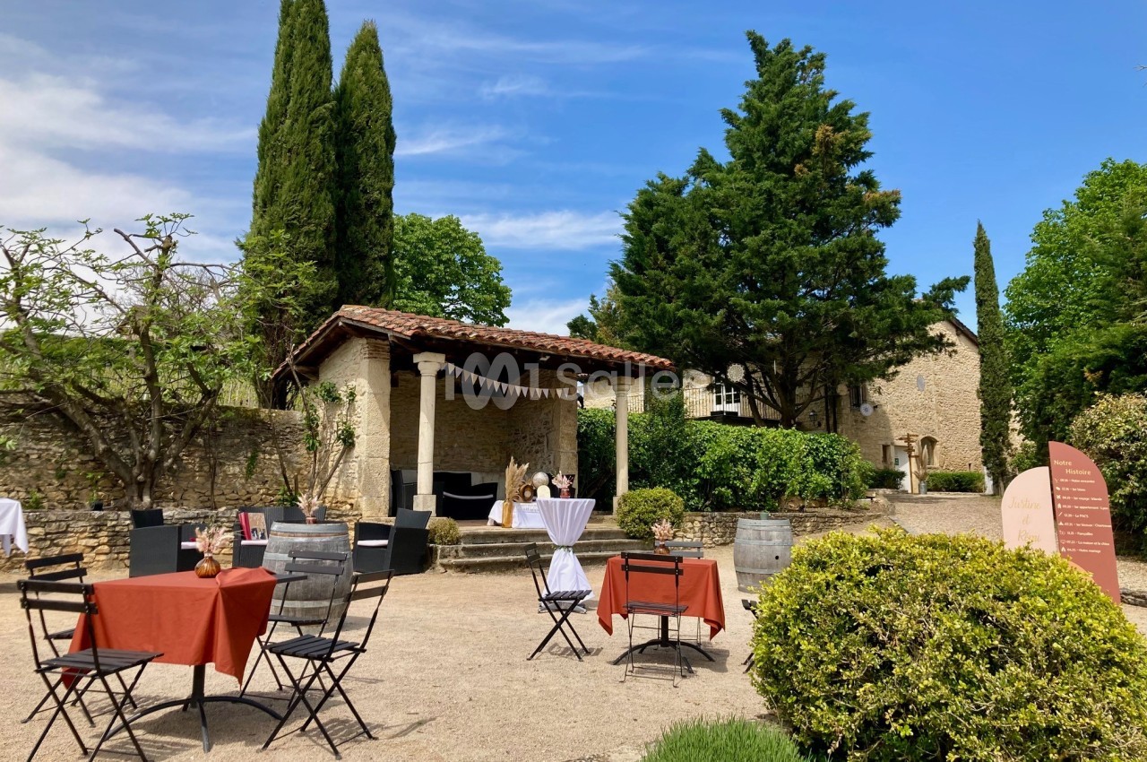 Terrasse extérieure avec tables dressées, pergola en pierre, arbres et bâtiments en pierre sous un ciel bleu.
