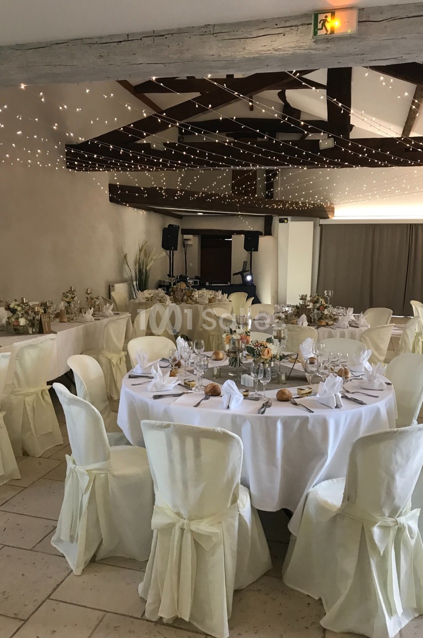 Salle de réception décorée avec des tables rondes dressées, nappes blanches et guirlandes lumineuses suspendues.