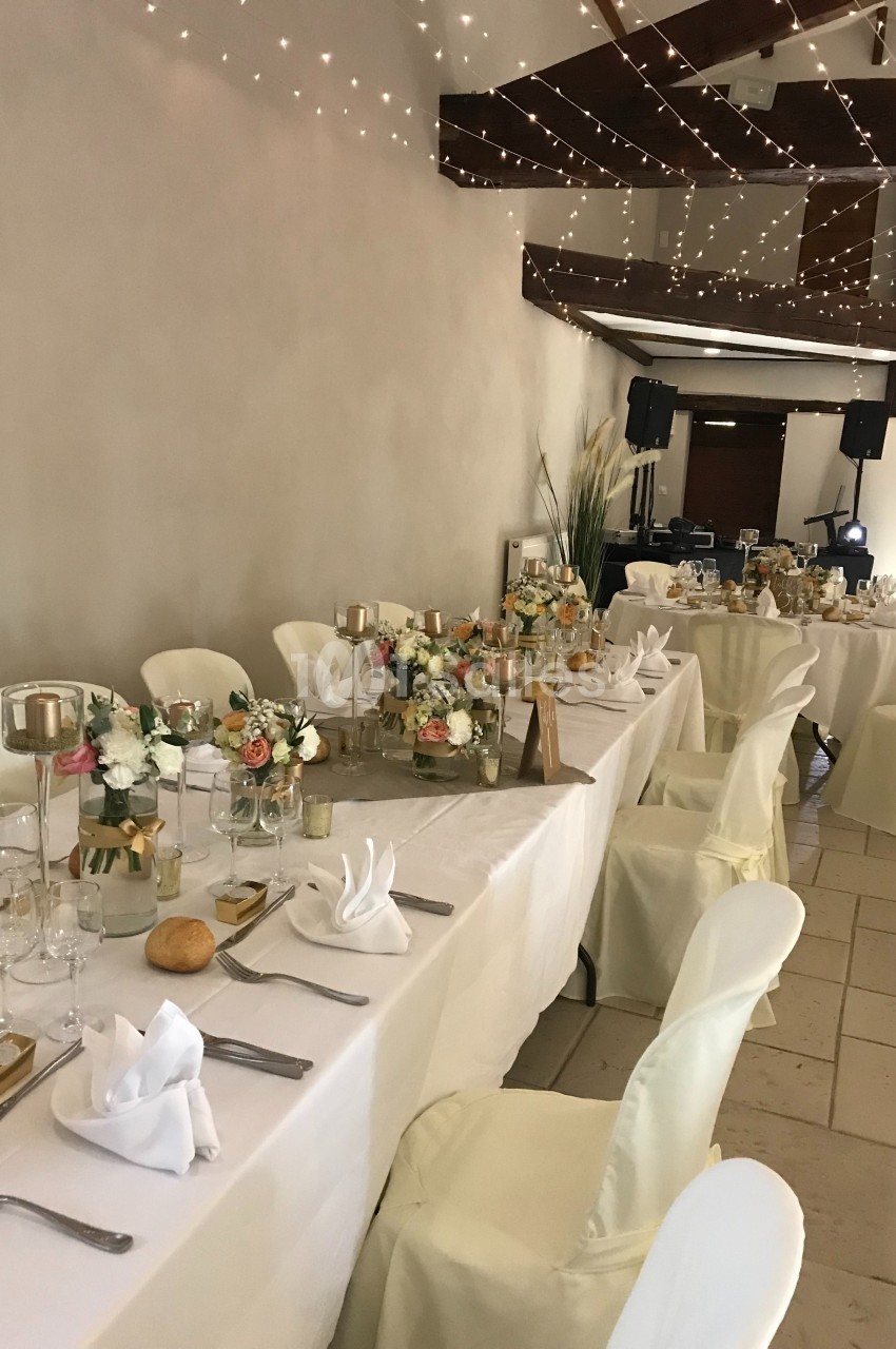 Salle de réception décorée avec des tables dressées, nappes blanches, arrangements floraux et guirlandes lumineuses…