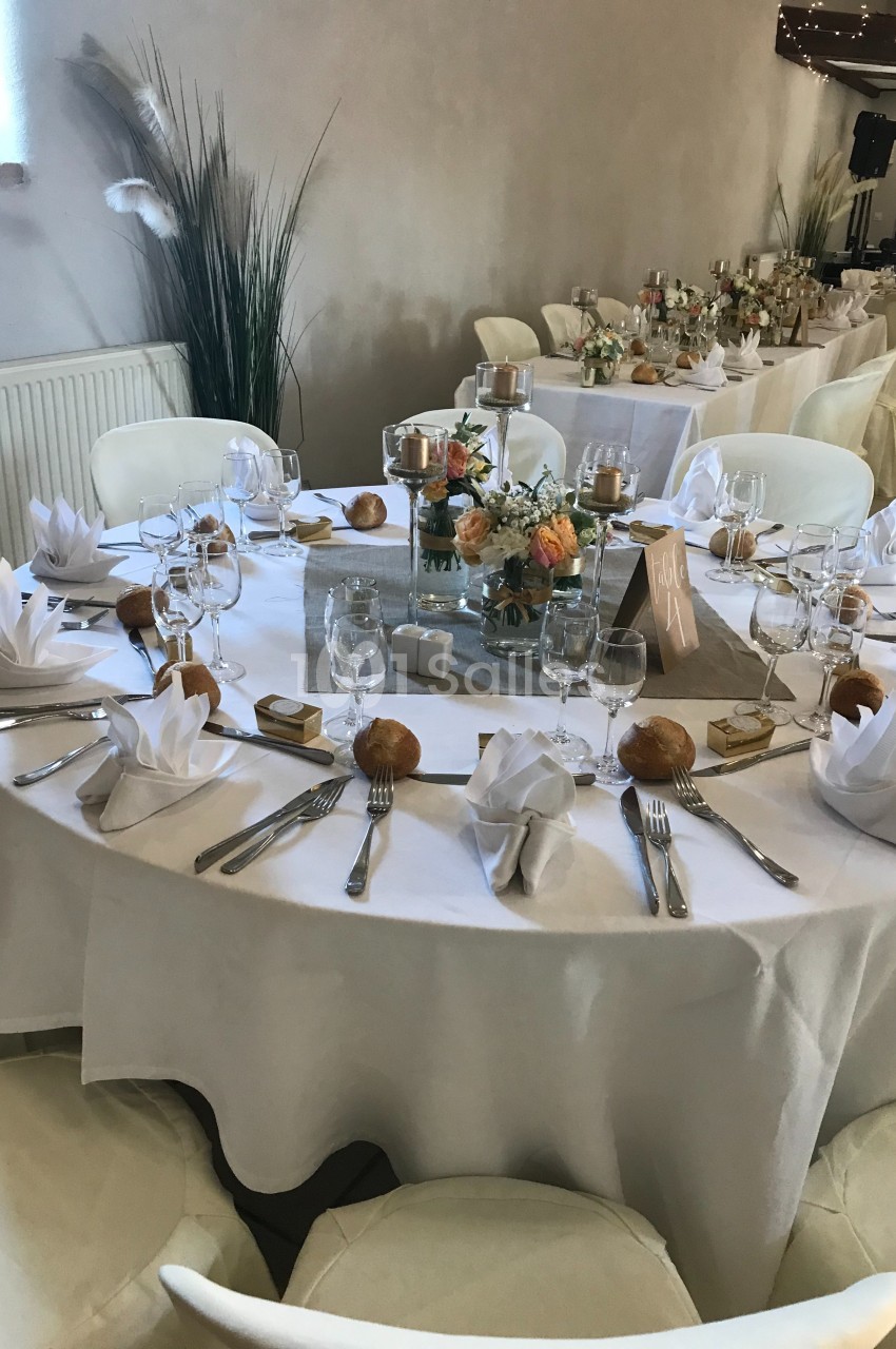 Table ronde dressée pour un événement, avec nappes blanches, serviettes pliées, vaisselle, bougies et décoration florale.