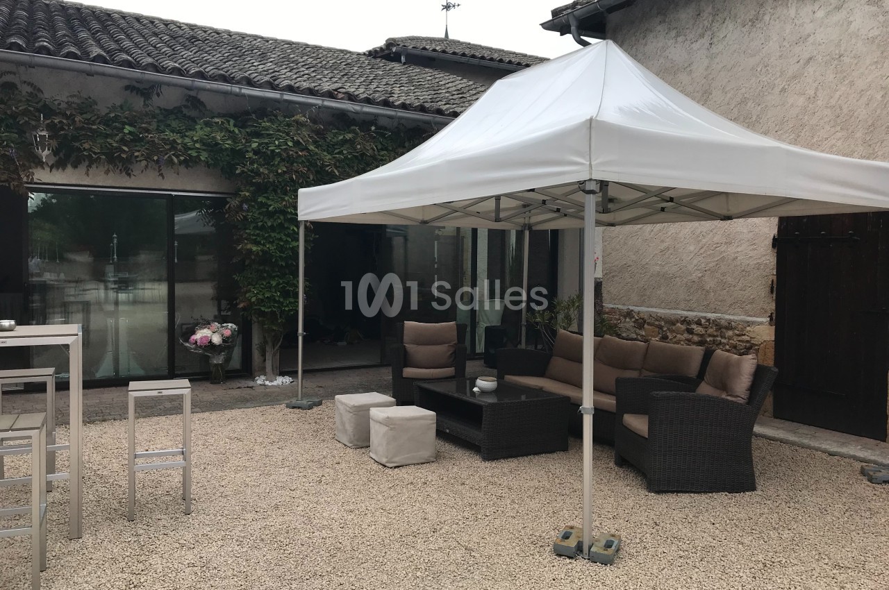 Tonnelle blanche installée dans une cour en gravier, avec salon de jardin en rotin et murs en pierre en arrière-plan.