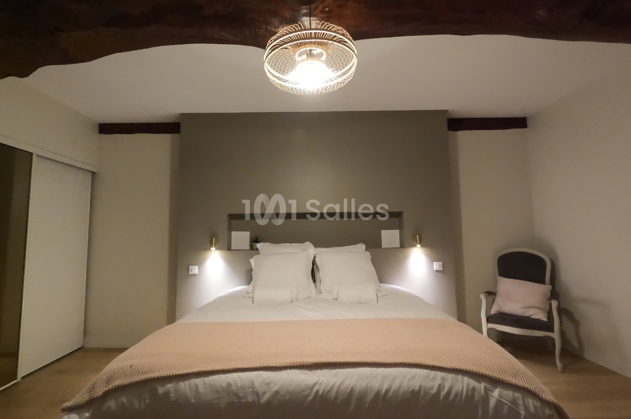 Lit double avec couverture beige dans une chambre lumineuse, éclairée par une suspension et décorée de poutres apparentes.