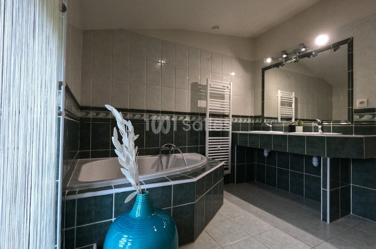 Salle de bain avec baignoire d'angle, carrelage vert et blanc, miroir éclairé et vase décoratif turquoise.