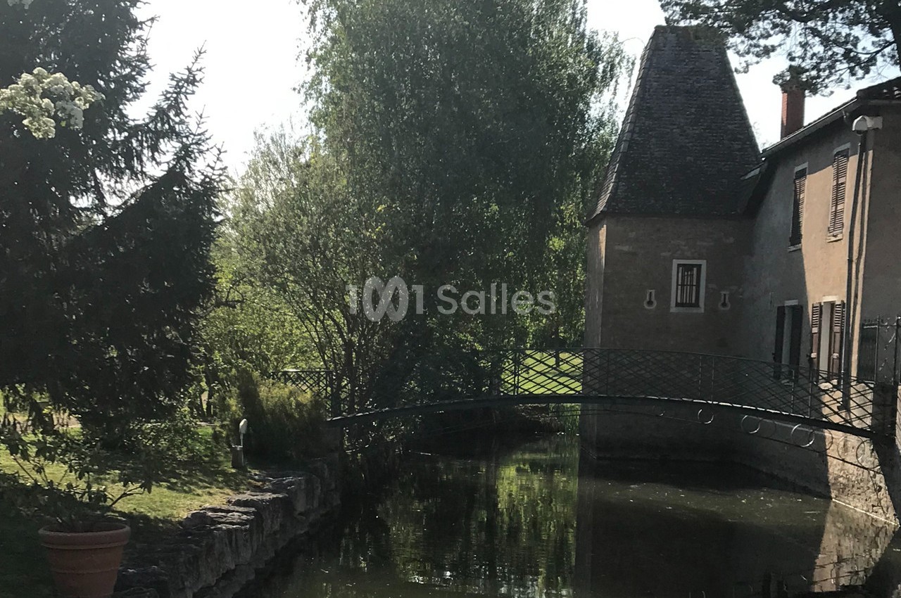 Petit pont en fer forgé traversant un cours d'eau bordé d'arbres, près d'une maison en pierre avec toit en ardoise.