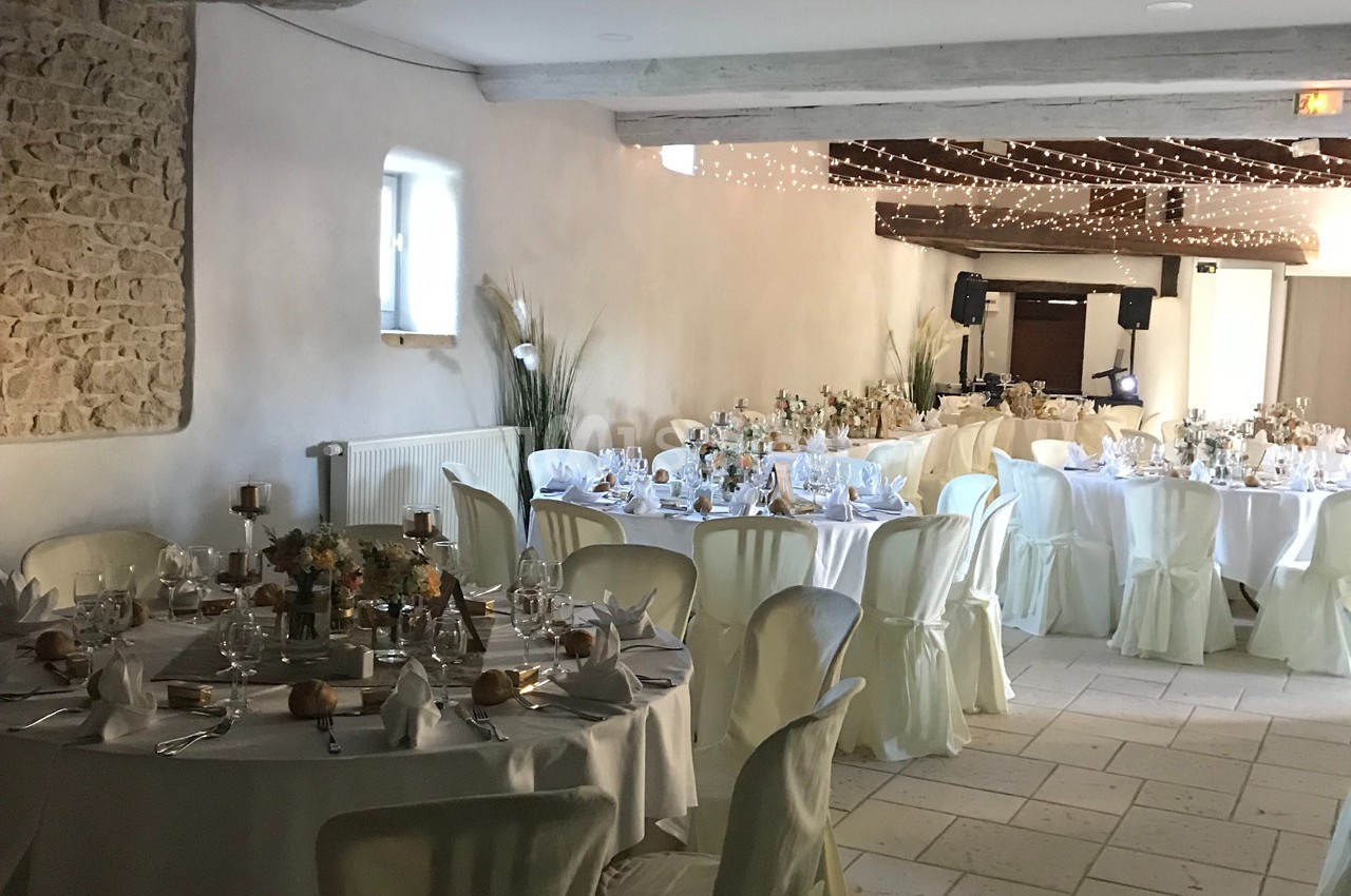 Salle de réception décorée pour un événement, avec tables rondes dressées, nappes blanches et guirlandes lumineuses au…
