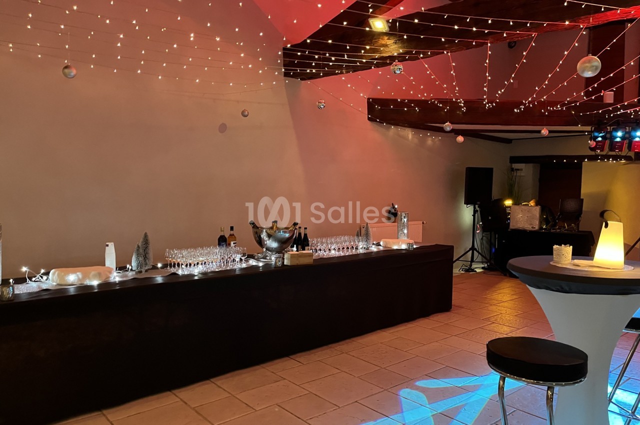 Salle décorée pour un événement, avec un bar dressé, des guirlandes lumineuses suspendues et une ambiance tamisée.
